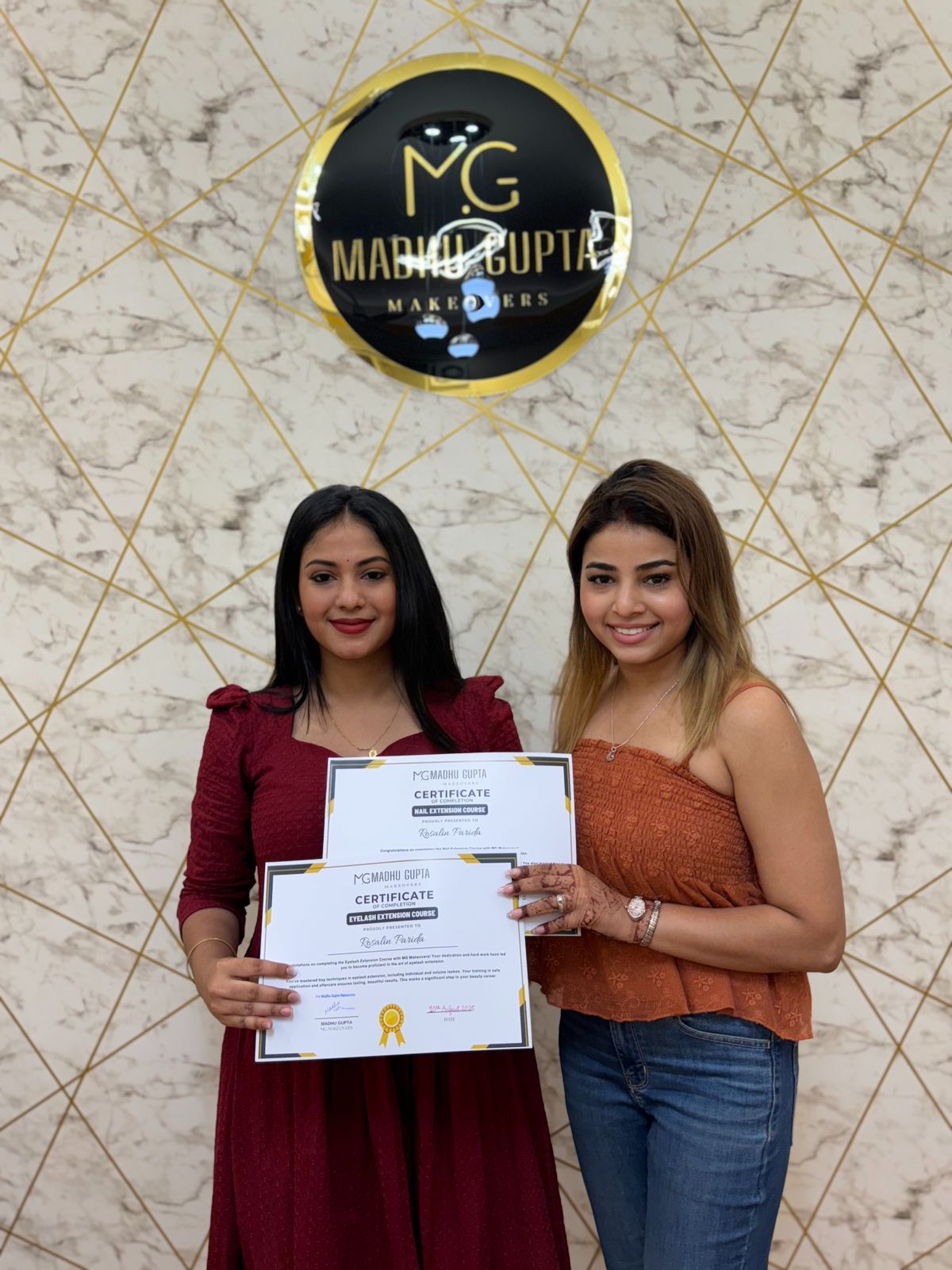 MG Makeovers Academy nail and lash graduates Two MG Makeovers Academy graduates holding Nail Extension and Eyelash Extension course certificates under the lit MG Madhu Gupta Makeovers logo