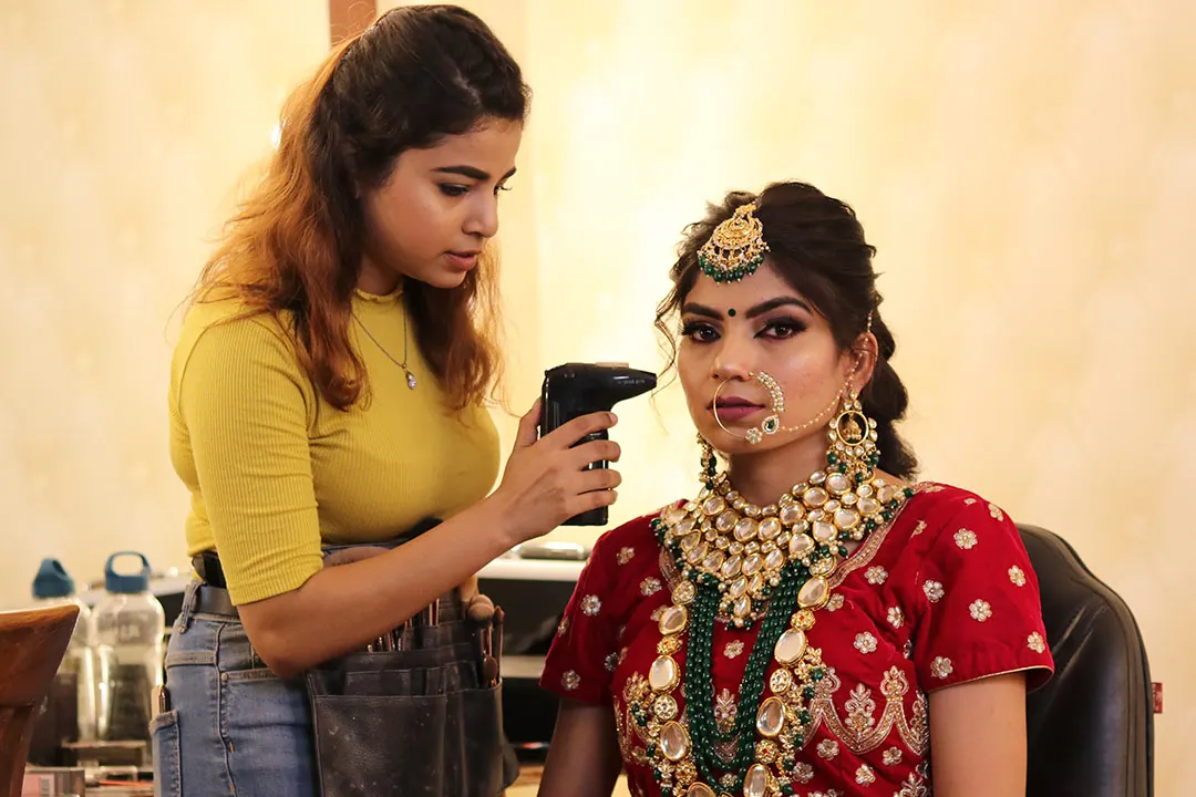 Airbrush bridal makeup by Madhu Gupta Madhu Gupta airbrushing makeup onto a bride in a red embroidered lehenga, kundan and emerald necklace, maang tikka and nath at MG Makeovers, Gurugram