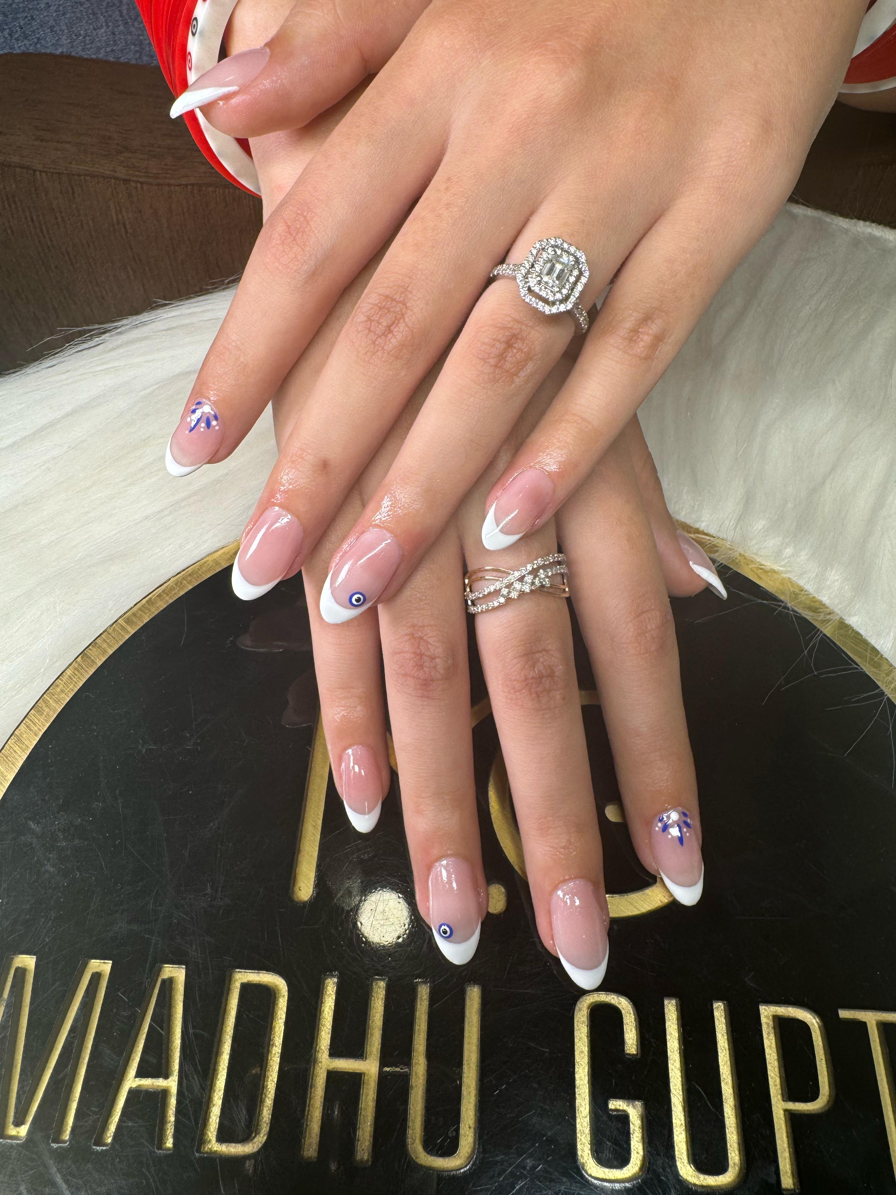 Evil eye French tip nails Almond nails with white French tips and small blue evil eye accents, a diamond ring on one hand, on the MG Makeovers logo plaque